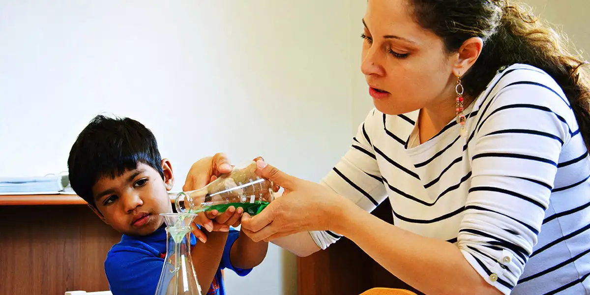 A teacher and a preschooler pouring liquid into a beaker together